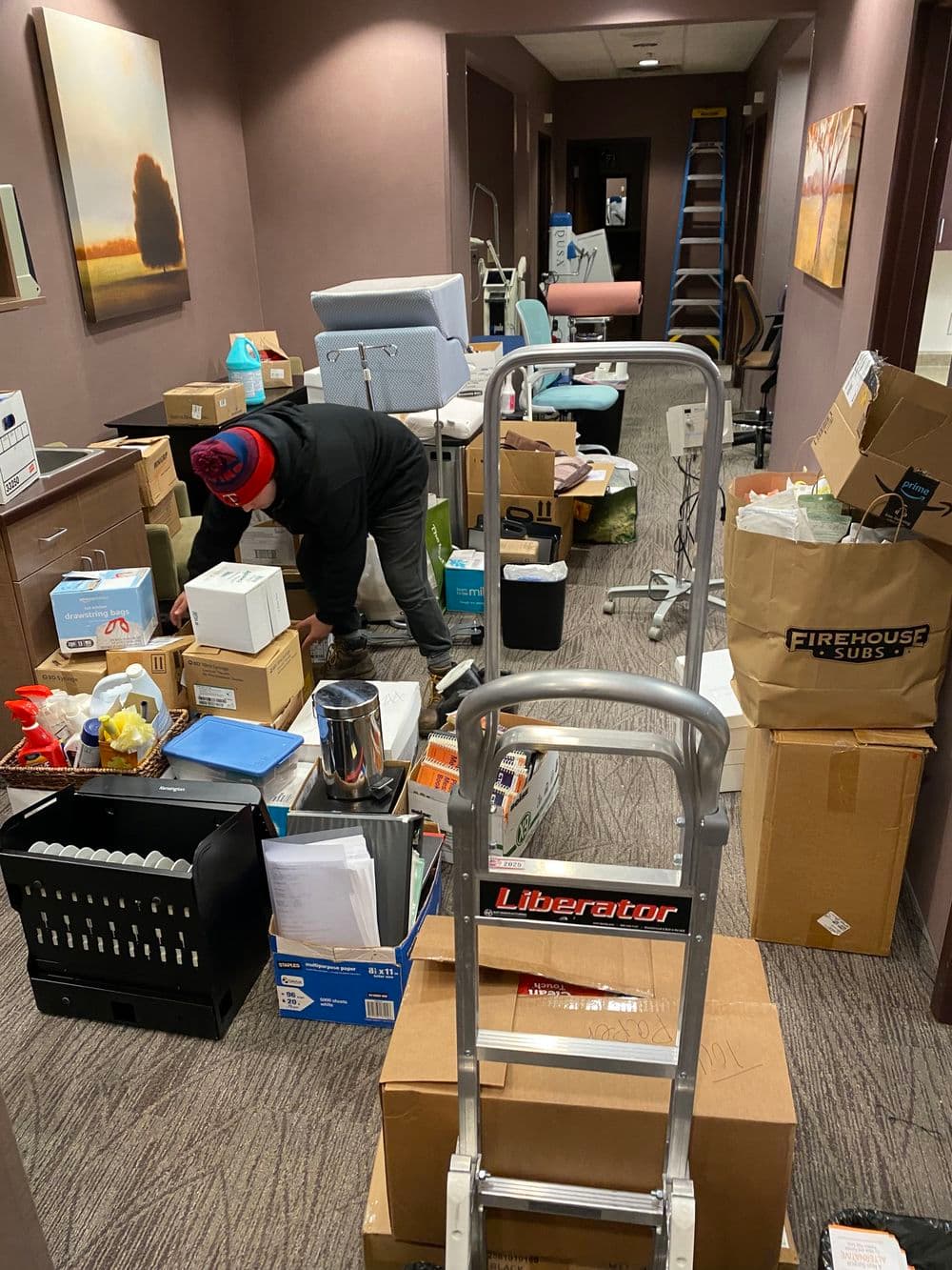 Person organizing cluttered hallway filled with boxes, office supplies, and furniture.
